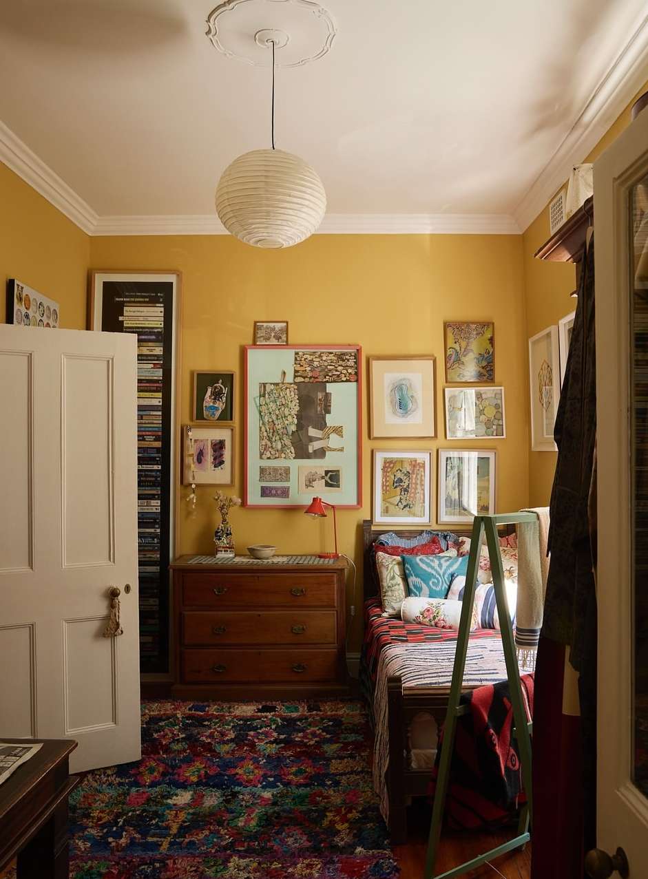 A warmly lit bedroom interior showcases a wooden bed, a chest of drawers, and vibrant artwork against yellow walls, under a paper lantern.