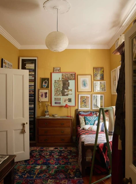 A warmly lit bedroom interior showcases a wooden bed, a chest of drawers, and vibrant artwork against yellow walls, under a paper lantern.