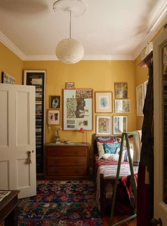 A warmly lit bedroom interior showcases a wooden bed, a chest of drawers, and vibrant artwork against yellow walls, under a paper lantern.