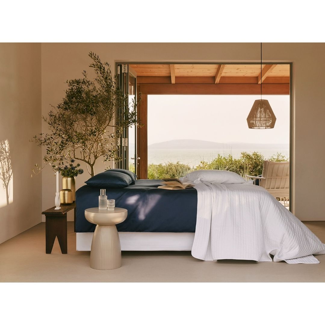 Serene interior showcases a styled bed with blue sheets and white quilt, complemented by natural light and decorative elements in a minimalist setting.