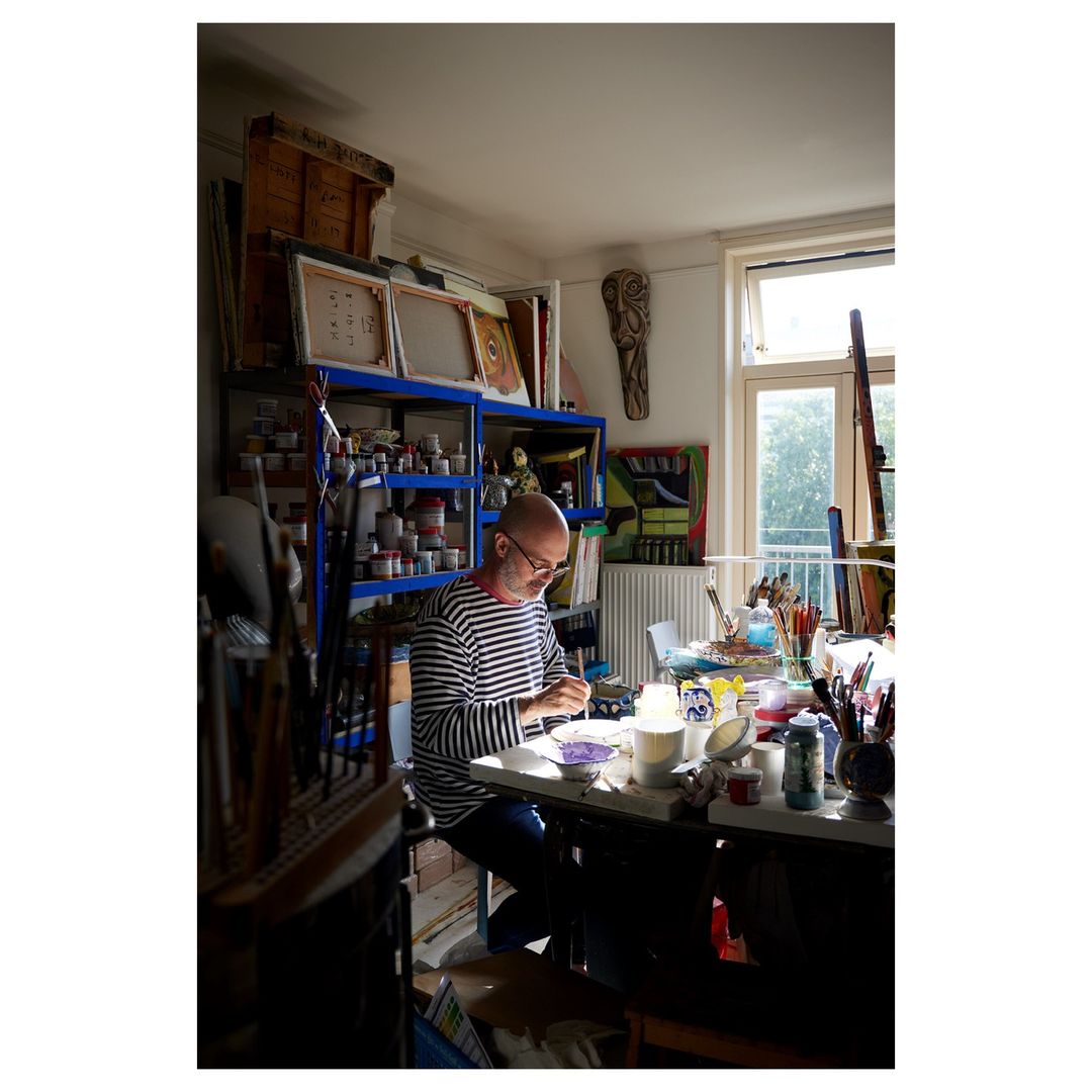Painter working in bright artist studio. Bald, bespectacled man in striped shirt sits at a cluttered table with art supplies.