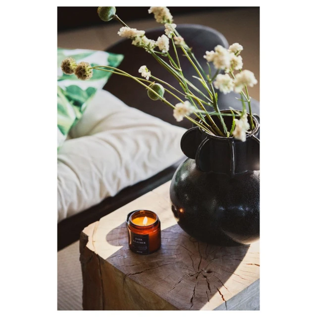 Still life featuring a black vase with white flowers, a lit candle, and a rustic wooden surface, set in an indoor environment with natural lighting.