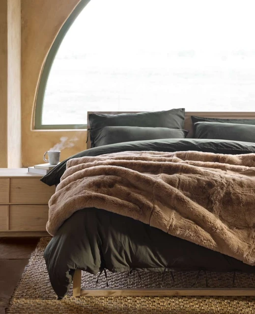 Cozy bedroom scene with a bed covered in a brown faux fur blanket, soft pillows, and a steaming mug on a bedside table.