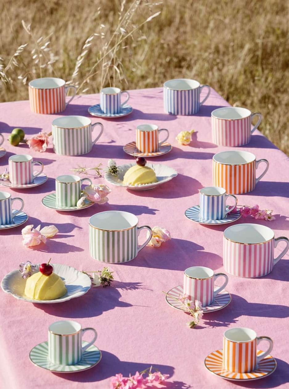 Striped teacups and saucers displayed on a pink tablecloth outdoors in a natural setting, arranged with desserts and flowers.