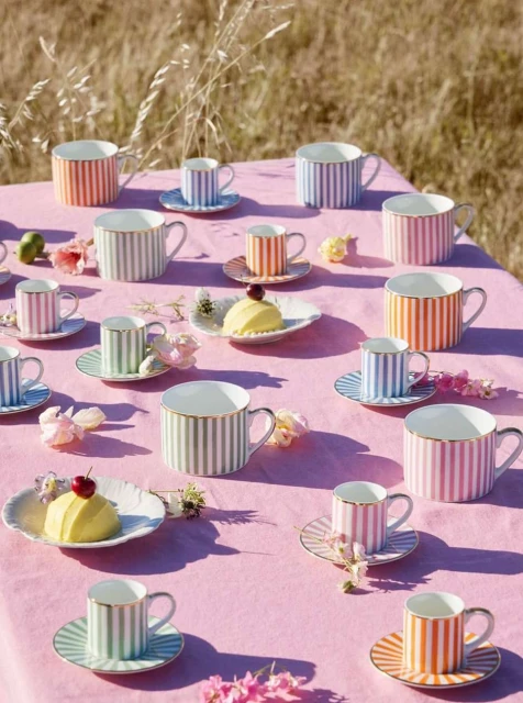 Striped teacups and saucers displayed on a pink tablecloth outdoors in a natural setting, arranged with desserts and flowers.