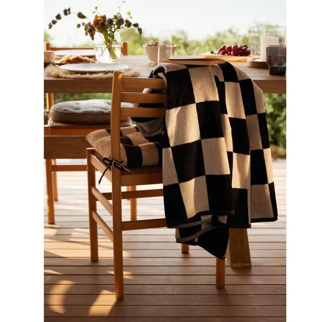 Wooden chair with checkered blanket and cushion near a wooden table with dishware and flowers in an outdoor dining setting.