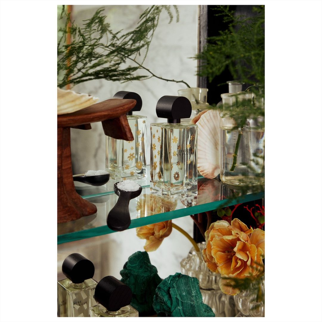 Still life of perfume bottles with white flowers, shells, and green plants on a glass shelf against a white marble backdrop.