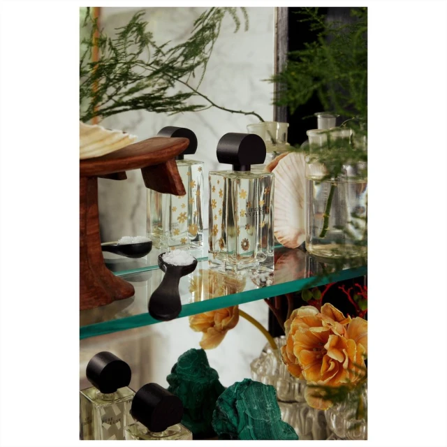 Still life of perfume bottles with white flowers, shells, and green plants on a glass shelf against a white marble backdrop.