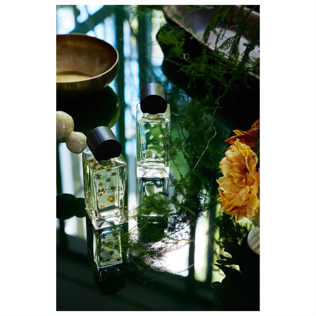 Two clear glass perfume bottles on a reflective surface surrounded by plants, flowers, and a bronze bowl in soft, natural light.