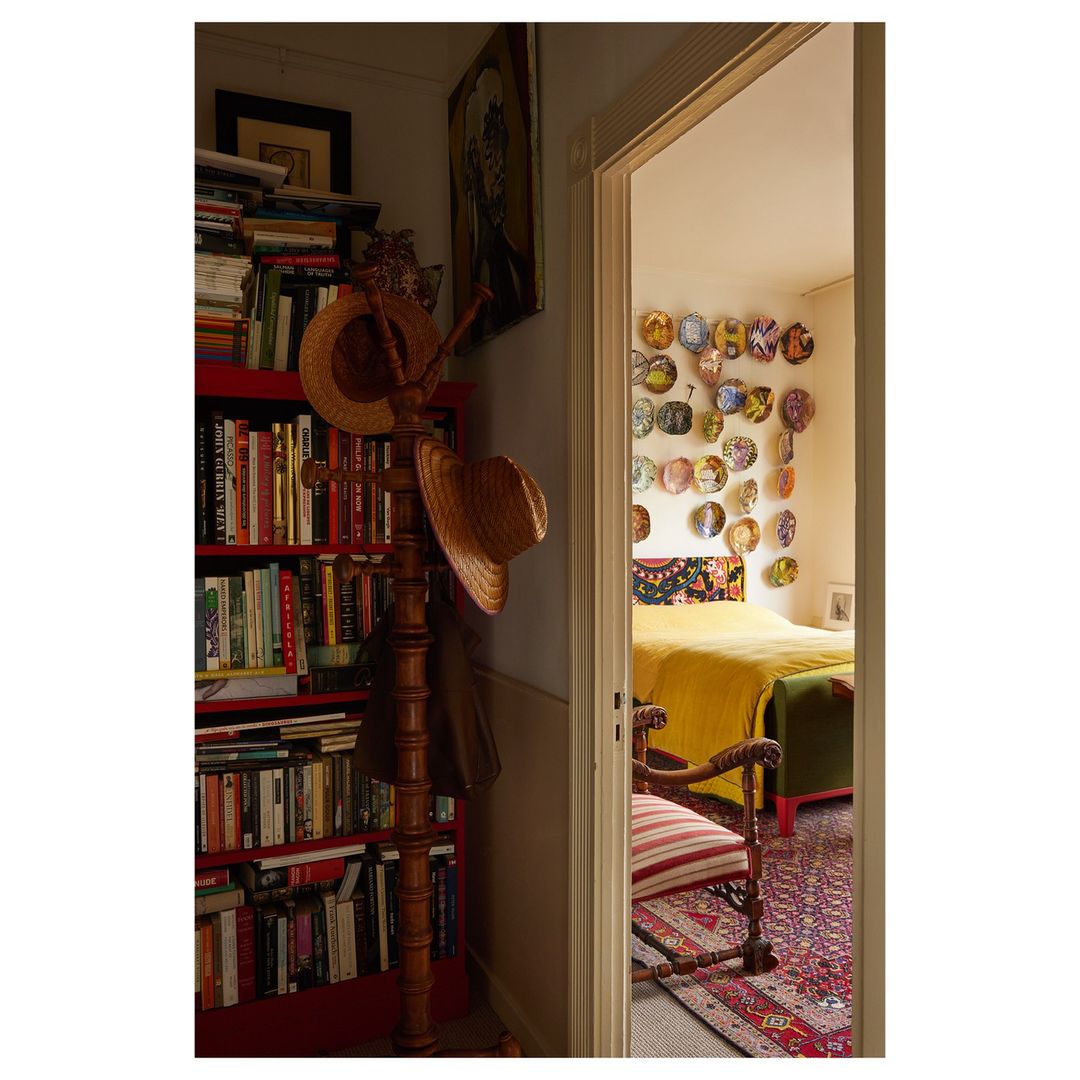 Cozy interior view through a doorway into a bedroom with a yellow bed, plates on the wall, with a bookcase and hat stand in the foreground.
