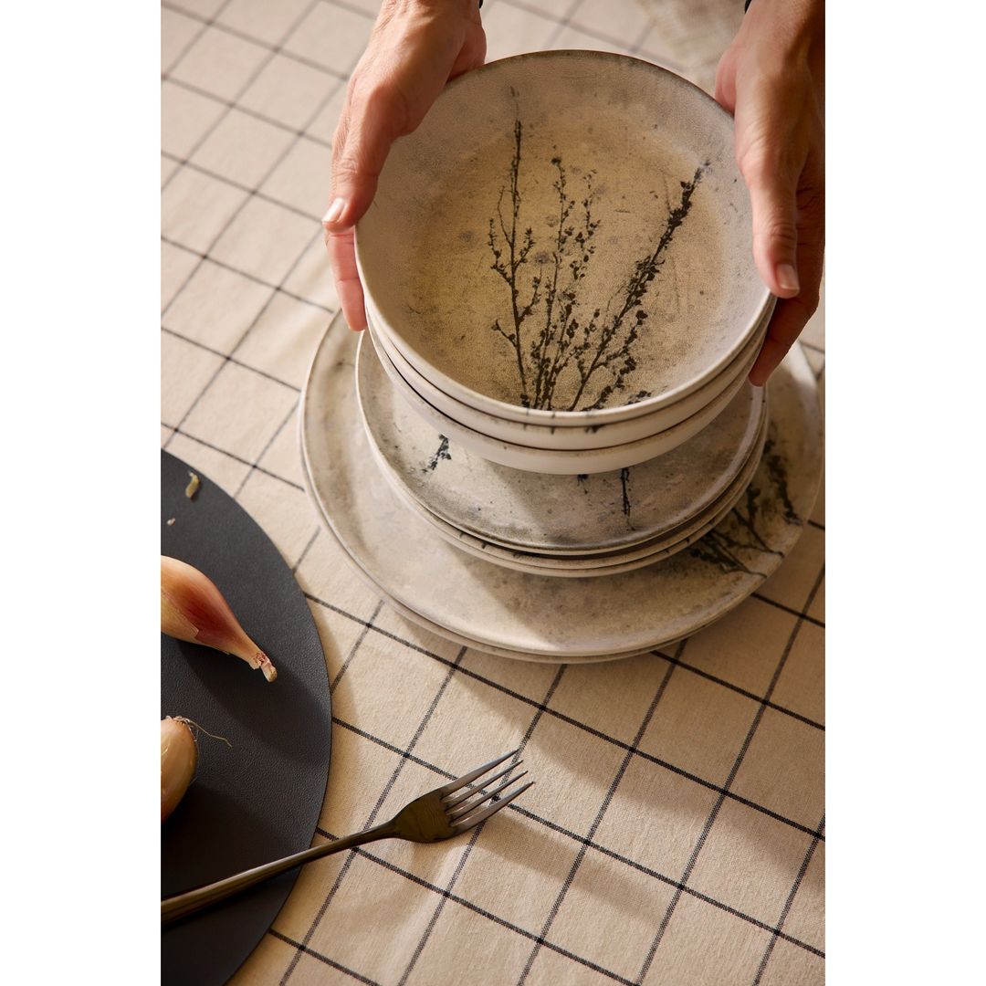 Close-up of hands arranging a stack of white ceramic plates with plant design on a plaid tablecloth beside a dark plate with shallots and a fork.