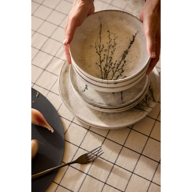 Close-up of hands arranging a stack of white ceramic plates with plant design on a plaid tablecloth beside a dark plate with shallots and a fork.