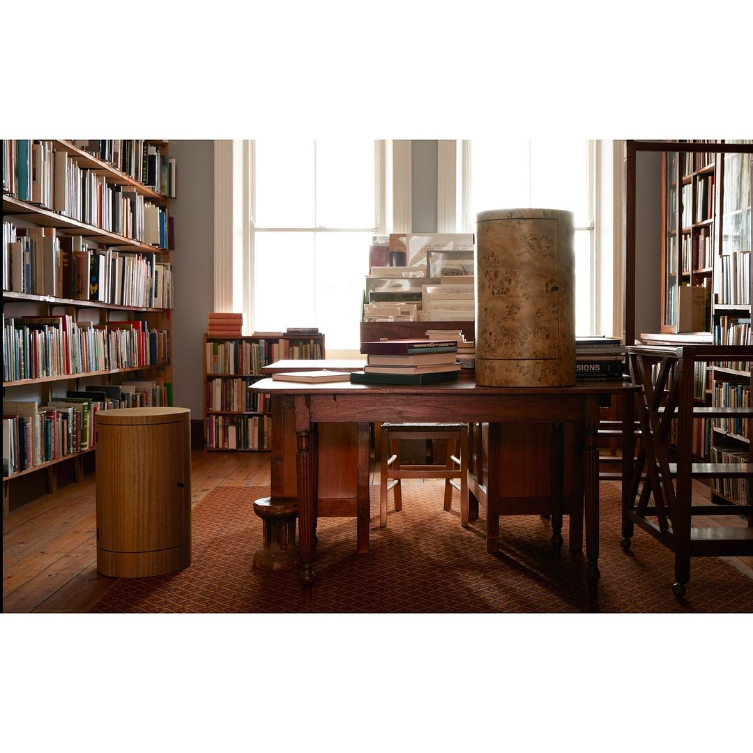 Interior of a library with wooden tables, chairs, bookcases filled with books, and natural light streaming through the windows.