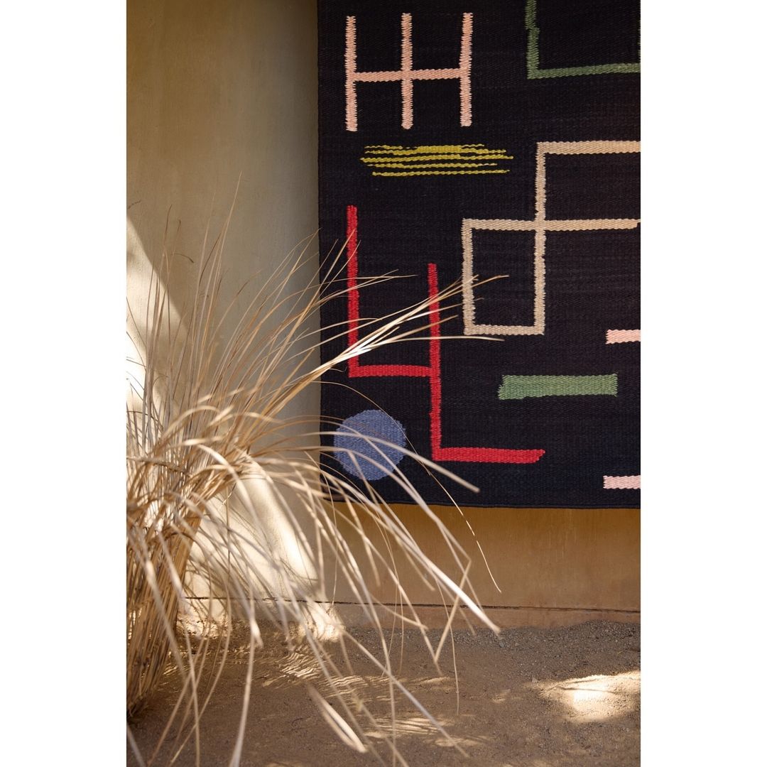 Abstract patterned wool rug hanging indoors with linear designs in coral, light blue, tan, green, and white on a dark background.
