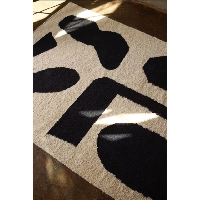 Beige wool rug with large black abstract shapes, laid on a concrete floor with natural light casting shadows across the textile.