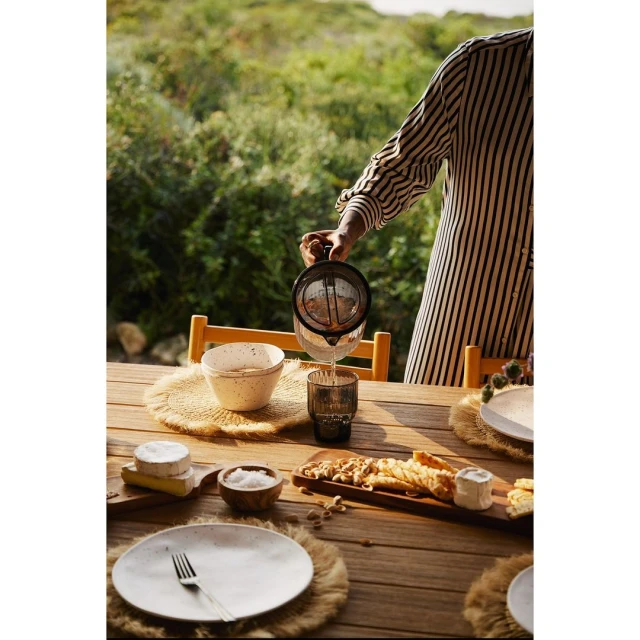 Person pours juice at an outdoor dining table featuring wooden furniture, plates, fruit, and greenery in the background.