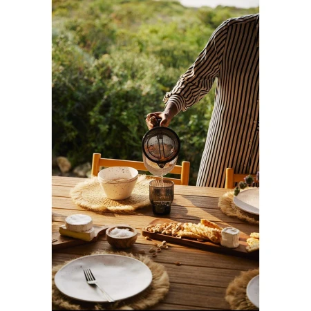 Person pours juice at an outdoor dining table featuring wooden furniture, plates, fruit, and greenery in the background.