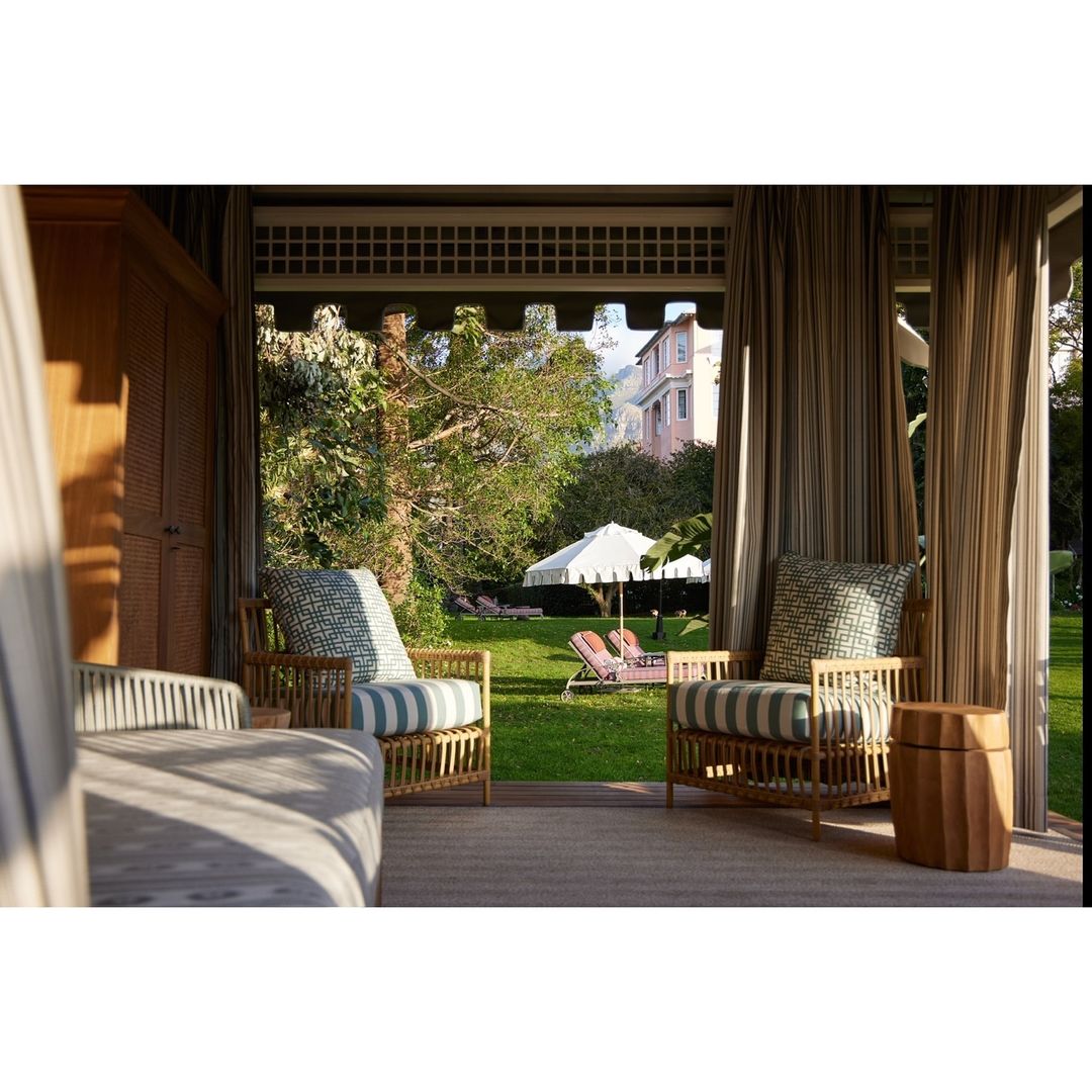 Outdoor lounge area with wicker chairs and striped cushions, featuring manicured lawn, garden foliage, and a glimpse of a building.