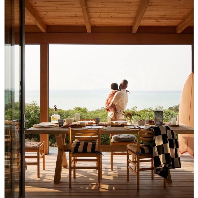 An adult carries a child on a deck overlooking a body of water. There is a dining table and chairs with a partial view of a surfboard.