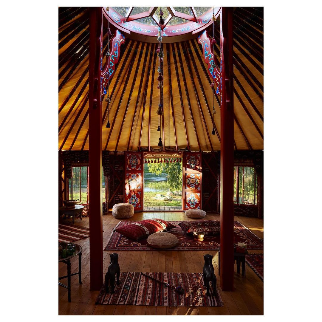 Interior view of a yurt-style dwelling with colorful textiles, pillows, rugs, and natural light from windows and a central skylight.