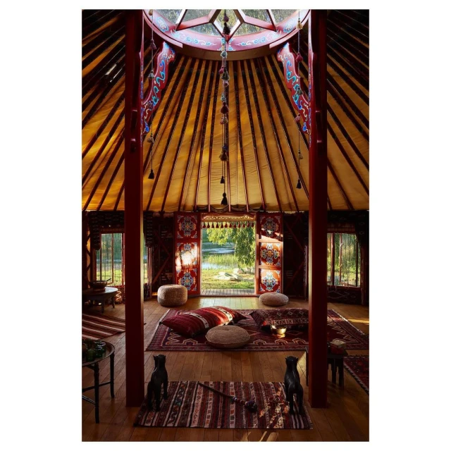 Interior view of a yurt-style dwelling with colorful textiles, pillows, rugs, and natural light from windows and a central skylight.