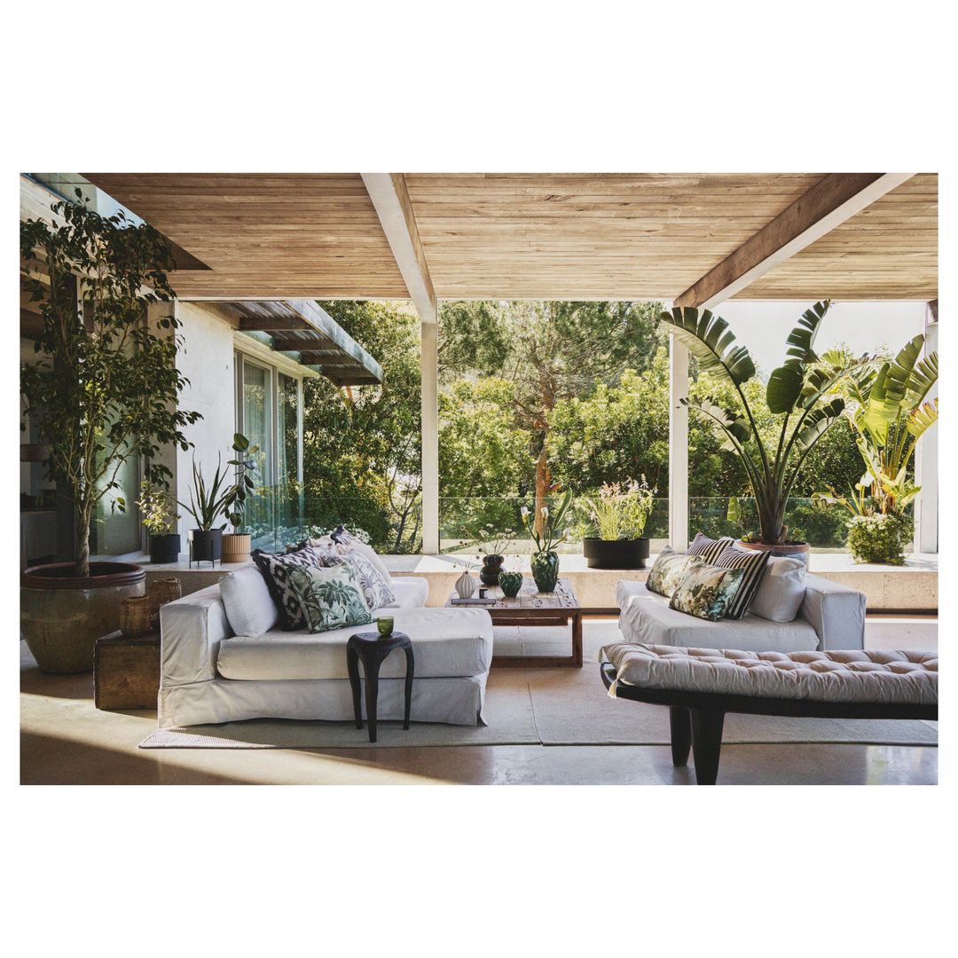 Sunlit veranda with a white slip-covered sofa, cushioned bench, and tropical plants, creating a relaxing indoor-outdoor living space.