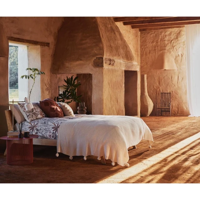 Bedroom features a bed with patterned linens, terra cotta colored walls, vase and chair, and light streaming from a window.