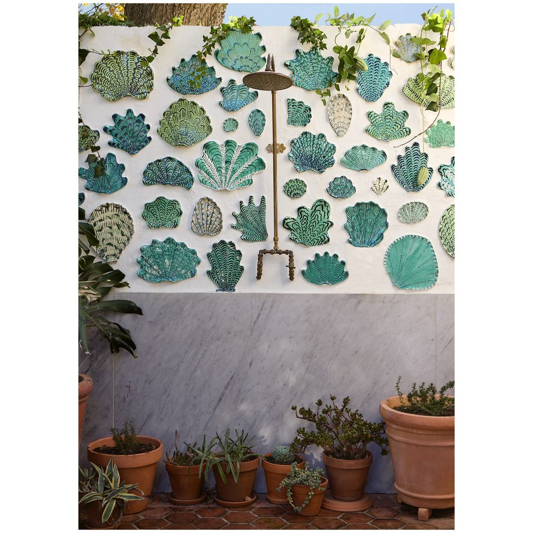 Outdoor shower fixture mounted on white wall with decorative seashell art, surrounded by potted plants and greenery, creating a backyard oasis.