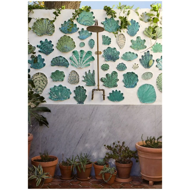 Outdoor shower fixture mounted on white wall with decorative seashell art, surrounded by potted plants and greenery, creating a backyard oasis.
