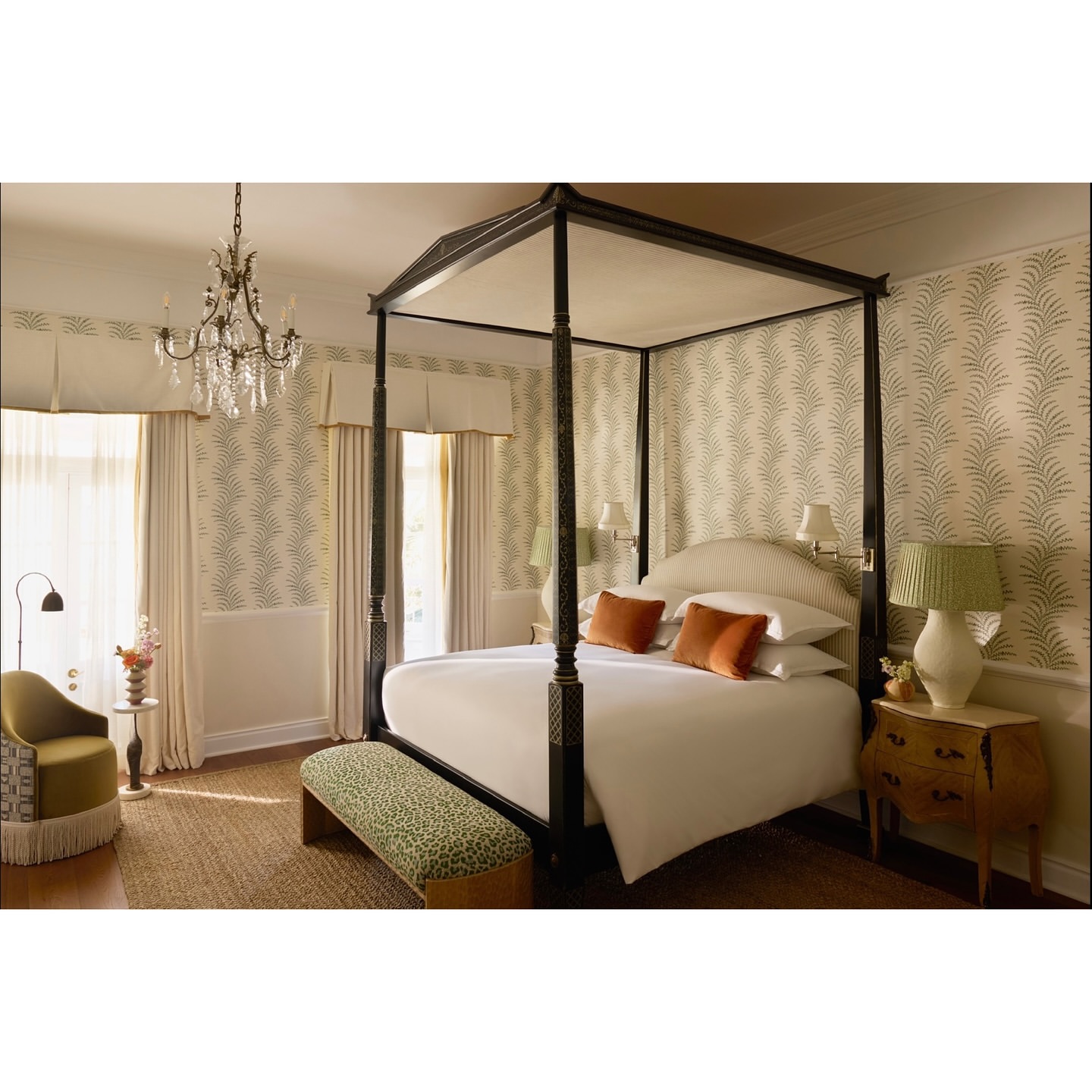 A four-poster bed in a hotel room is the main focus, showcasing an opulent interior with patterned wallpaper, chandelier, and stylish furniture.