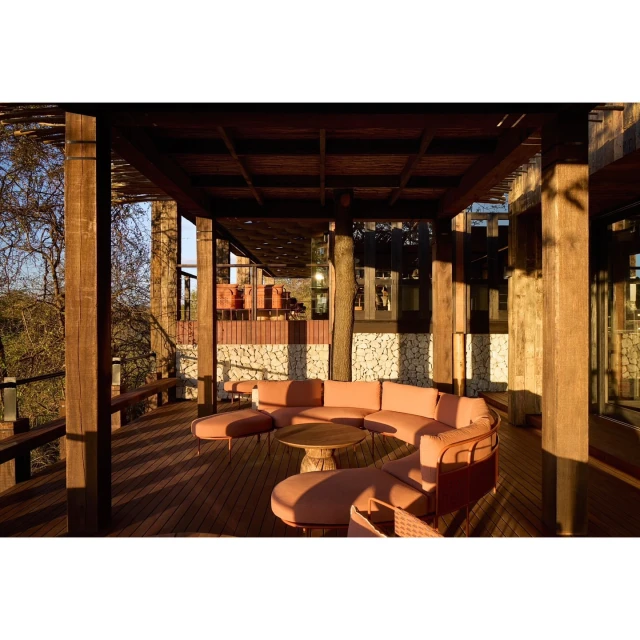 Outdoor seating area with a coral-colored sectional sofa and a wooden table on a wooden deck, surrounded by trees and wooden architectural details.