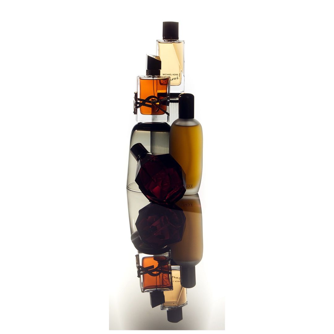 Five assorted perfume bottles are stacked vertically, featuring various shapes and colors, against a stark white background with a reflective surface.