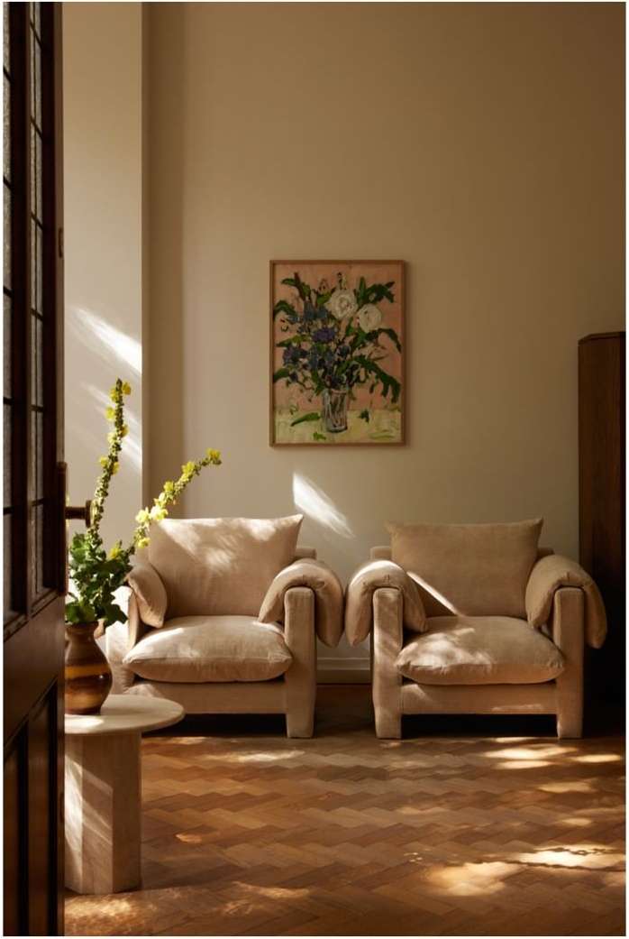 Interior photo of a living room with two beige armchairs, wooden herringbone floor, and a floral painting above the armchairs. 