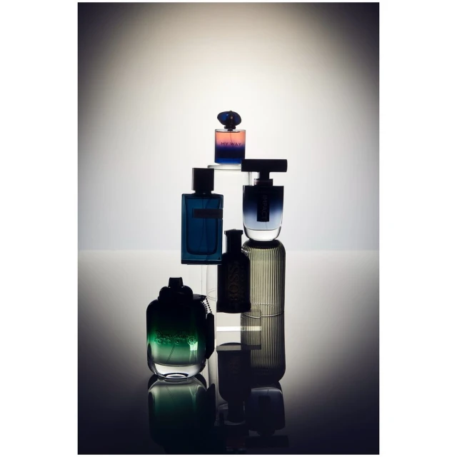 Still life of multiple assorted perfume bottles in shades of blue, green, and violet arranged on clear acrylic risers in studio lighting.