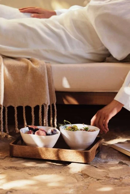 Person reclining on bed with wooden tray holding two white bowls filled with snacks. Natural light and minimalist design.
