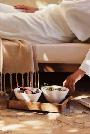 Person reclining on bed with wooden tray holding two white bowls filled with snacks. Natural light and minimalist design.
