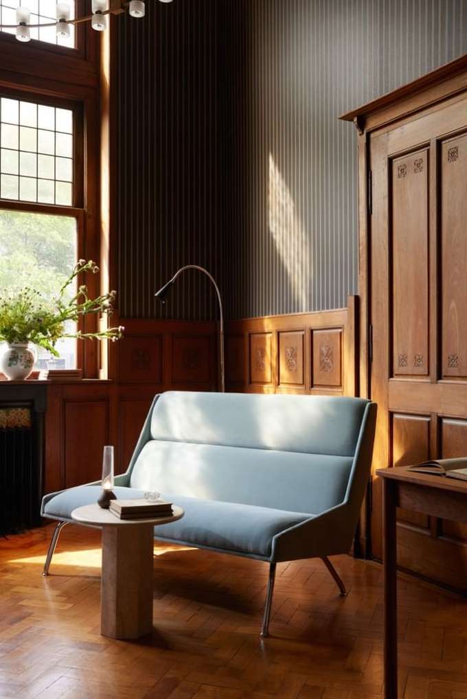 Modern light blue sofa with a small round table and books sits on a wooden floor in a naturally lit room with striped wallpaper.