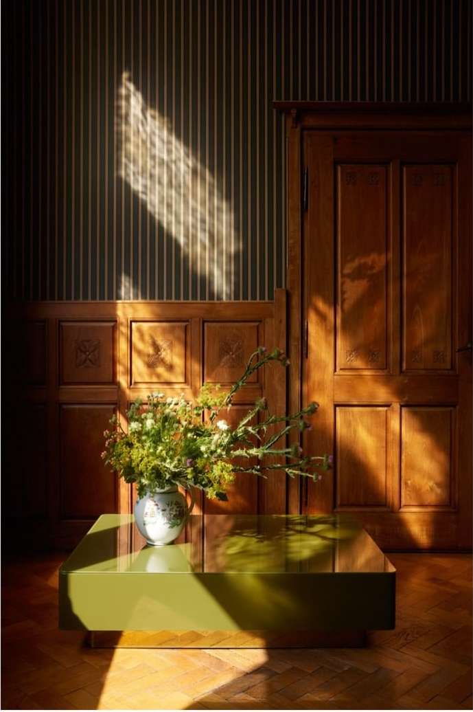 Still life of a green table with flowers in a vase in a room with striped wallpaper, creating a warm and inviting ambiance.