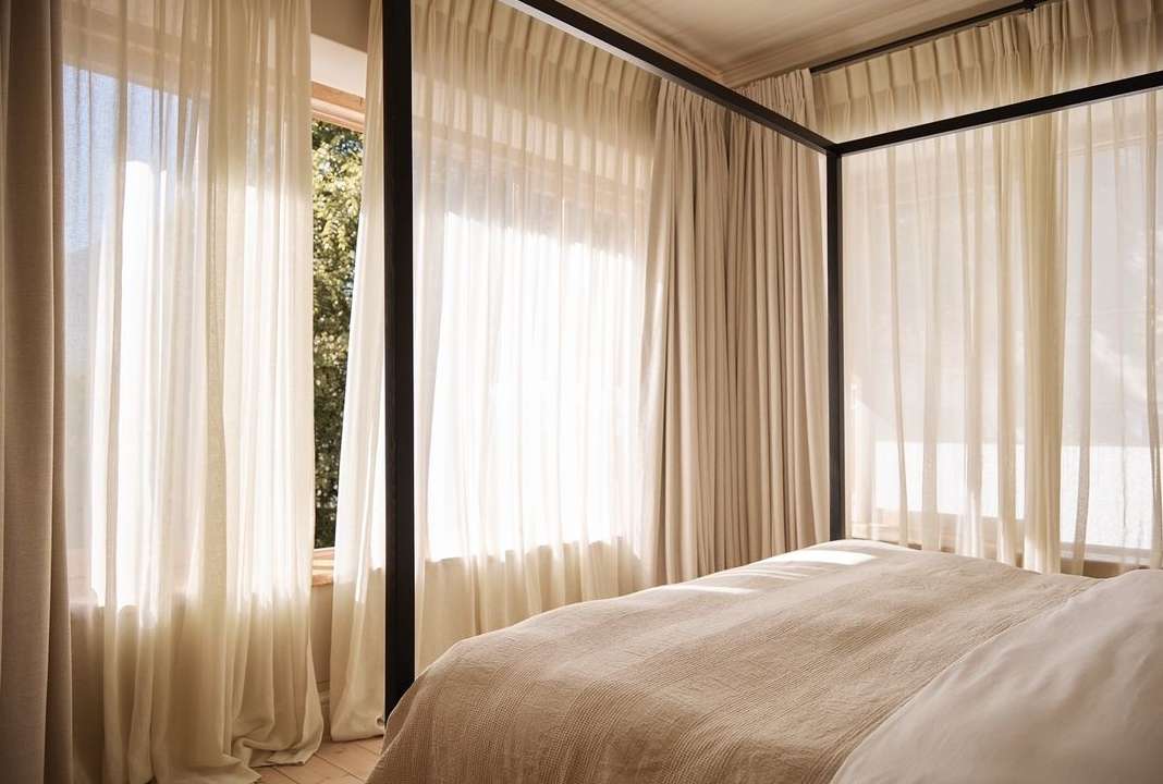 Interior of a bedroom featuring a canopy bed with light-colored bedding and sheer curtains filtering sunlight through a window.
