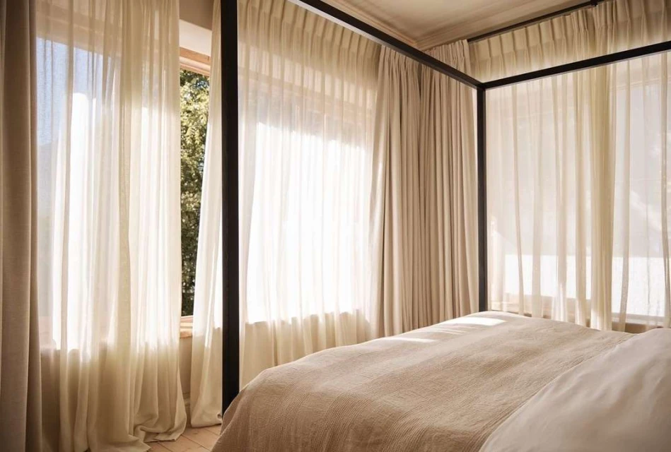 Interior of a bedroom featuring a canopy bed with light-colored bedding and sheer curtains filtering sunlight through a window.