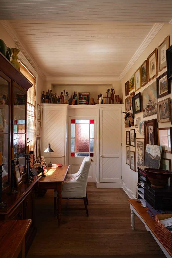 Home office interior with wood desk, white chair, gallery wall of framed art, and collectibles on top of built-in cabinets.