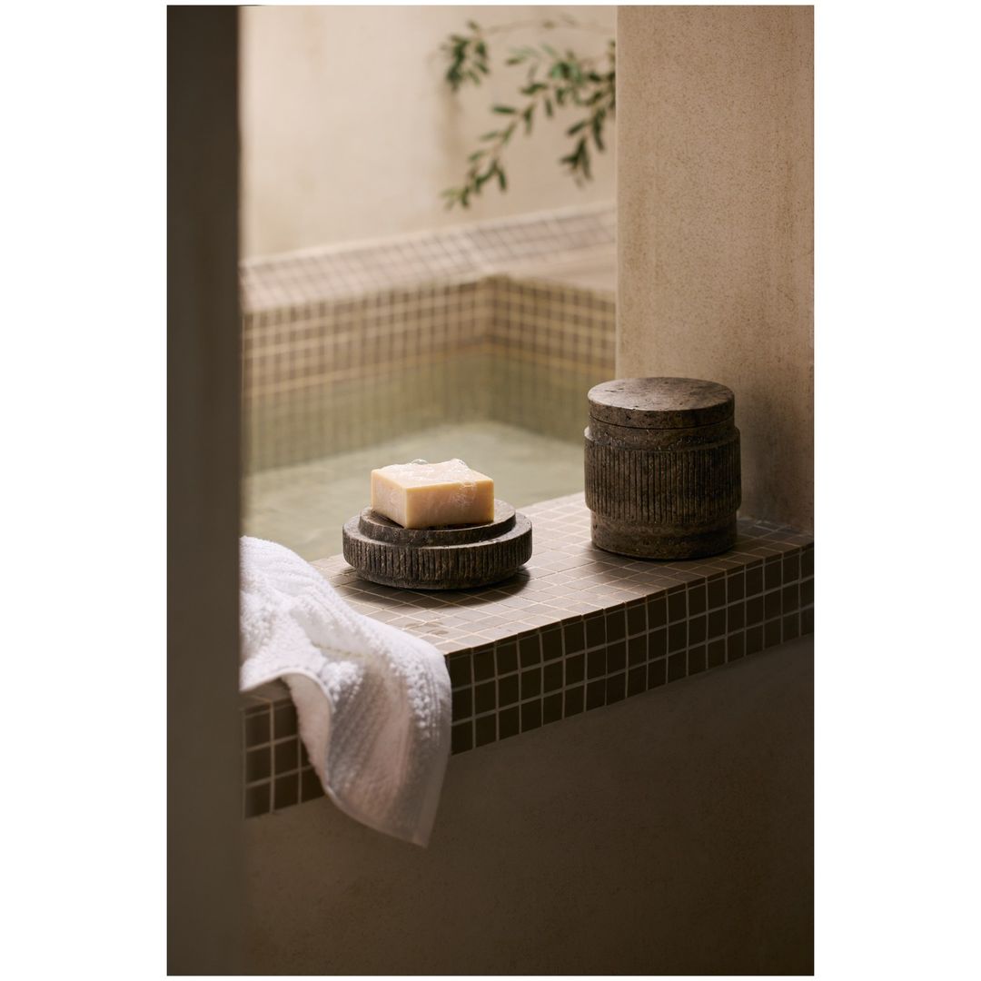 Still life featuring a soap bar on a dark, carved dish and a decorative container, staged in a bathroom setting with a tiled backdrop.
