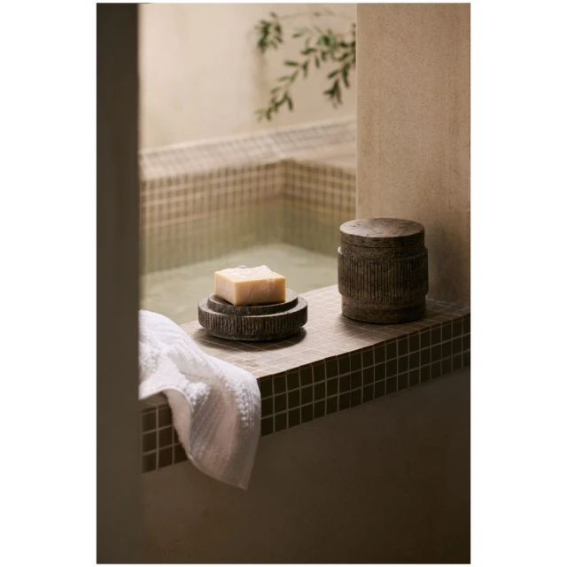 Still life featuring a soap bar on a dark, carved dish and a decorative container, staged in a bathroom setting with a tiled backdrop.