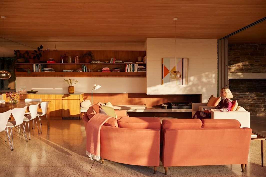 Spacious modern living room interior with a salmon-colored couch, dining table, blond wood paneling, and a woman sitting on a white sofa.