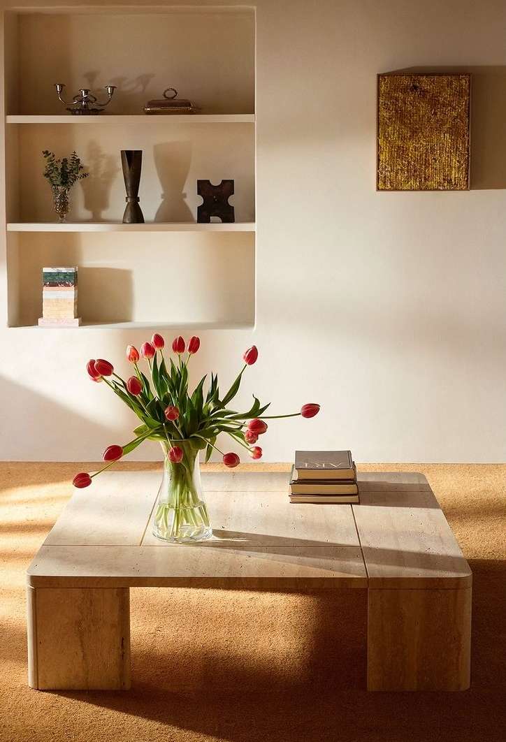 Interior shot of a vase with red tulips on a travertine coffee table in front of a shelving unit with decorative objects.