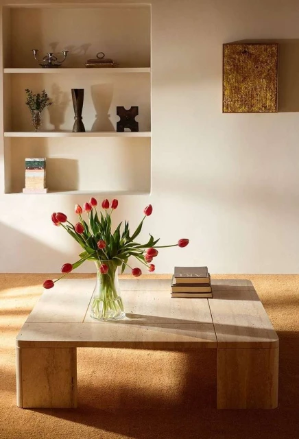 Interior shot of a vase with red tulips on a travertine coffee table in front of a shelving unit with decorative objects.