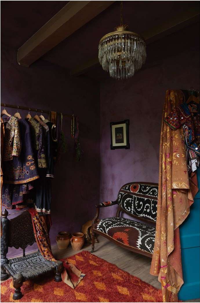 Interior of room with decorative clothing rack, patterned chair, orange rug, and hanging chandelier; purple walls in background.