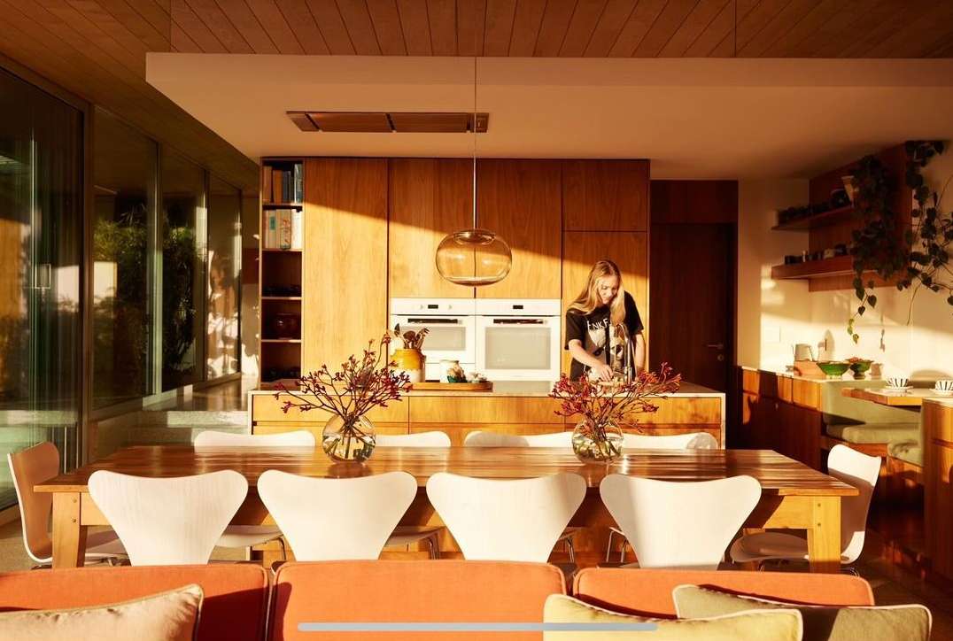 Sunlit modern home interior with a woman standing at a kitchen island, wooden cabinets, a dining table, and orange upholstered seating.