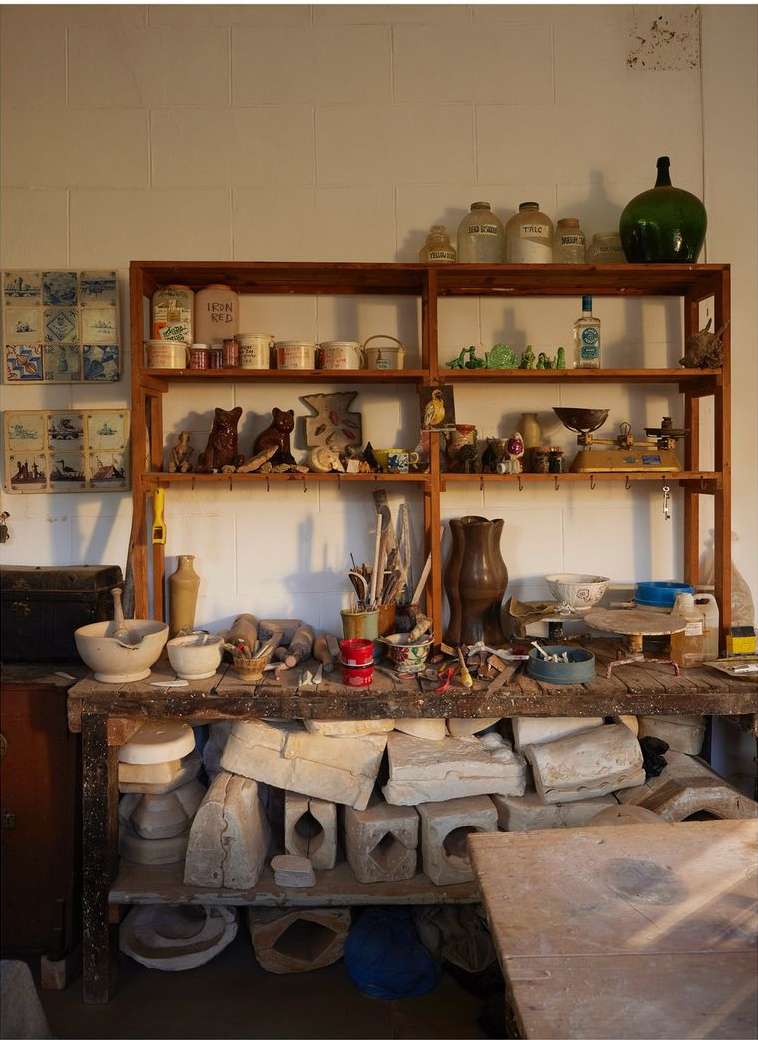 Indoor still life of a workbench covered with plaster molds, paint brushes, and ceramic tools, surrounded by shelves with vintage bottles and jars.