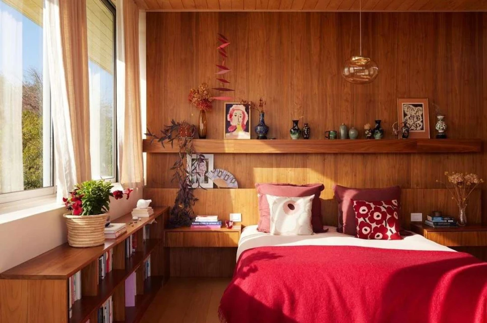 Bedroom interior with mid-century modern decor, featuring a red bedspread, wood-paneled walls, and natural light from a window.